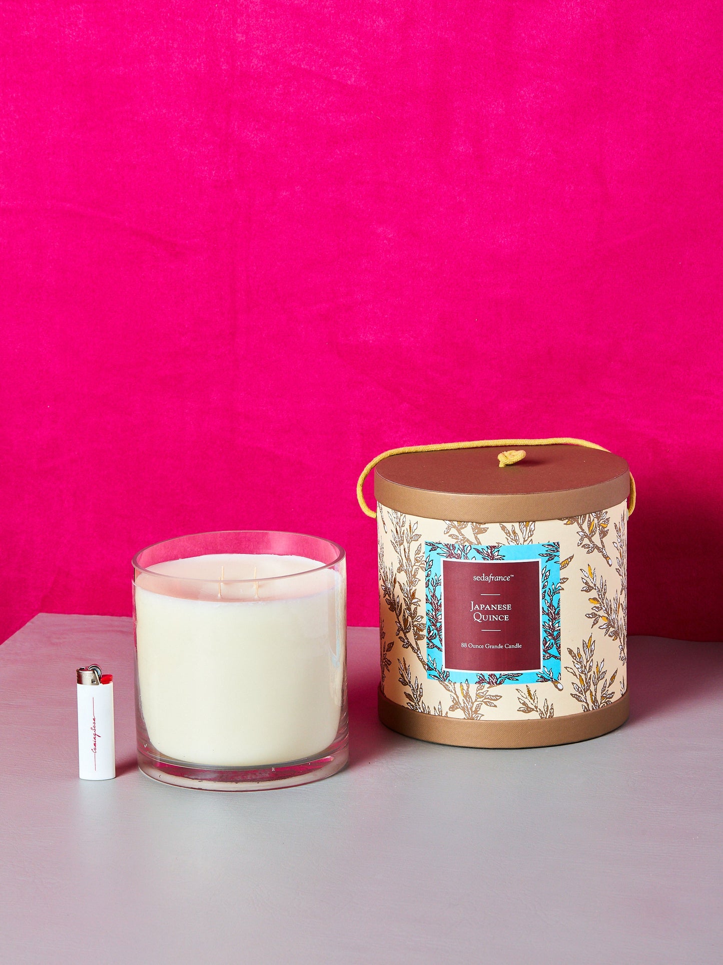 A large 88 ounce Japanese Quince candle sits besides its cylindrical decorative box and a white Coming Soon lighter.